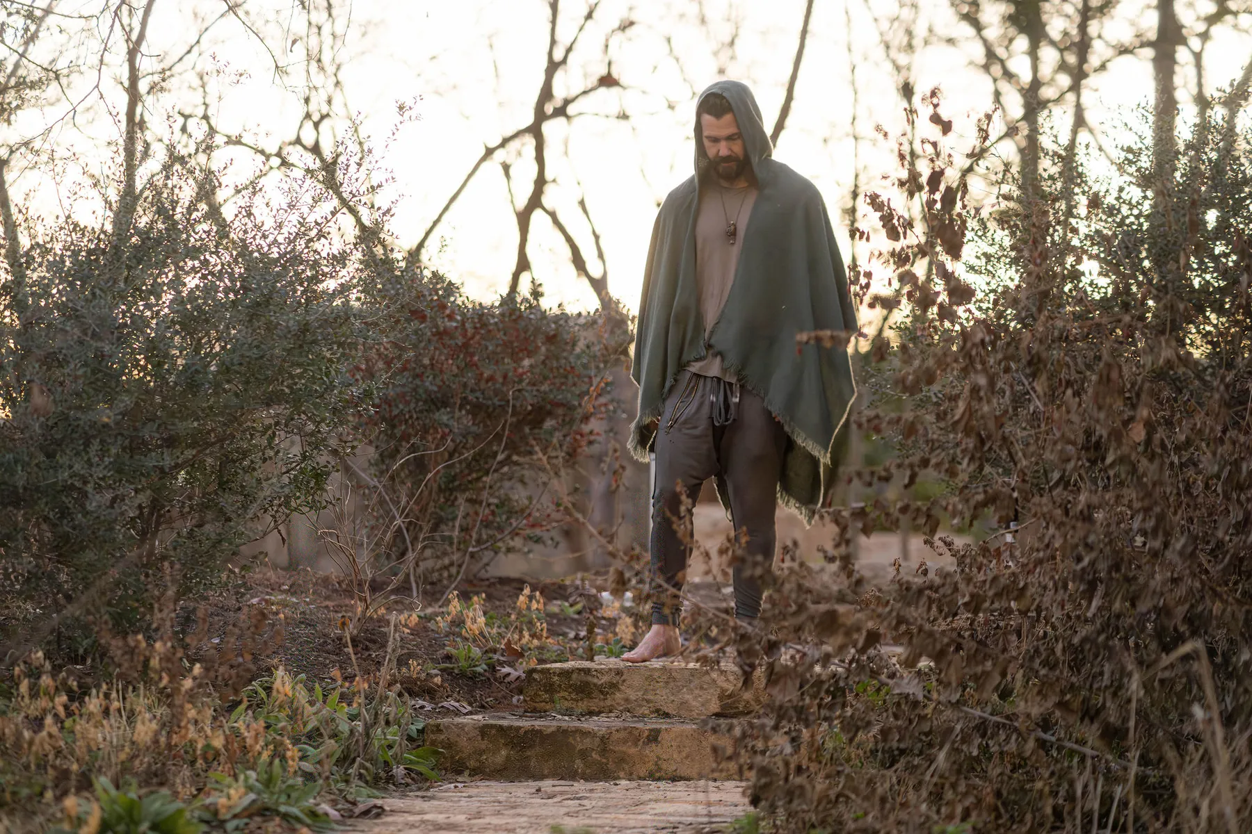 Zack Blakeney walking barefoot down stone steps in a winter garden, wrapped in a hooded cloak.